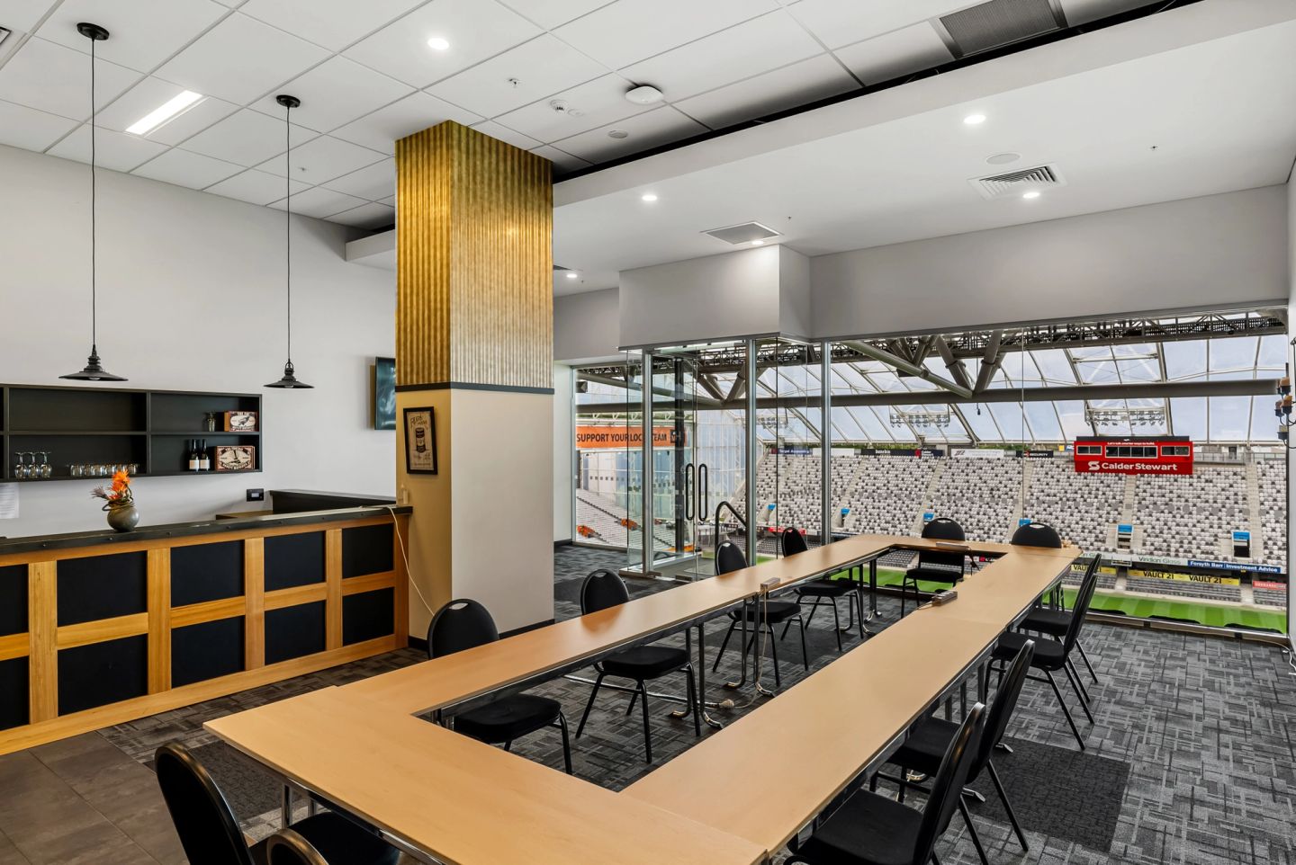 Small meeting room with U-shaped table, black chairs, a bar counter, and glass wall overlooking stadium field