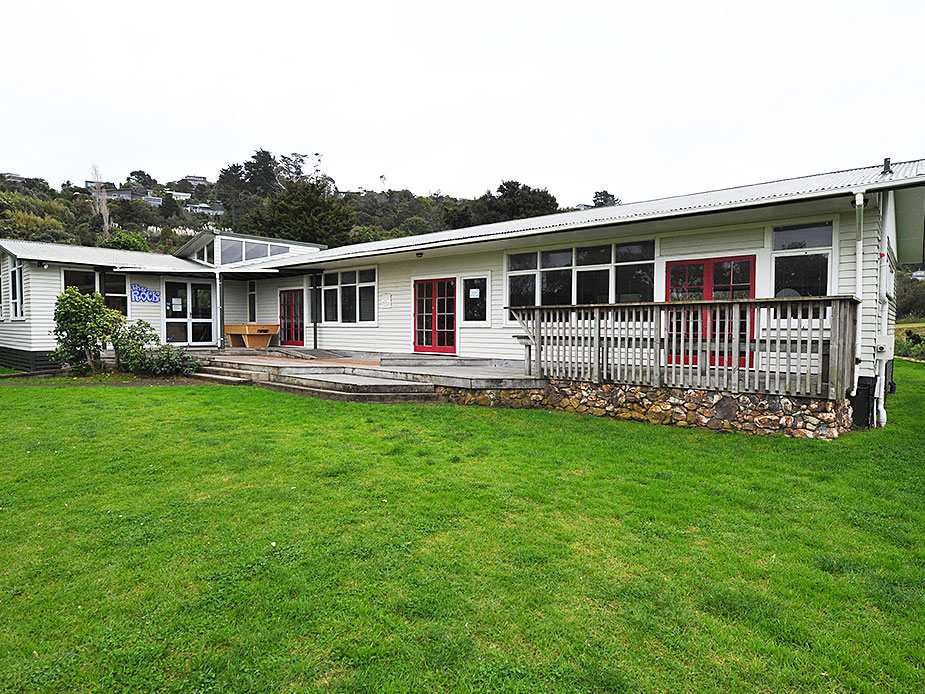 White weatherboard hall with red window frames, veranda, and lawn