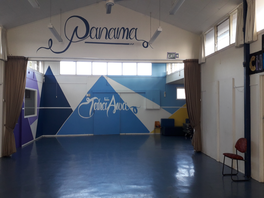 Single-storey blue community hall building with mural, fence and car park