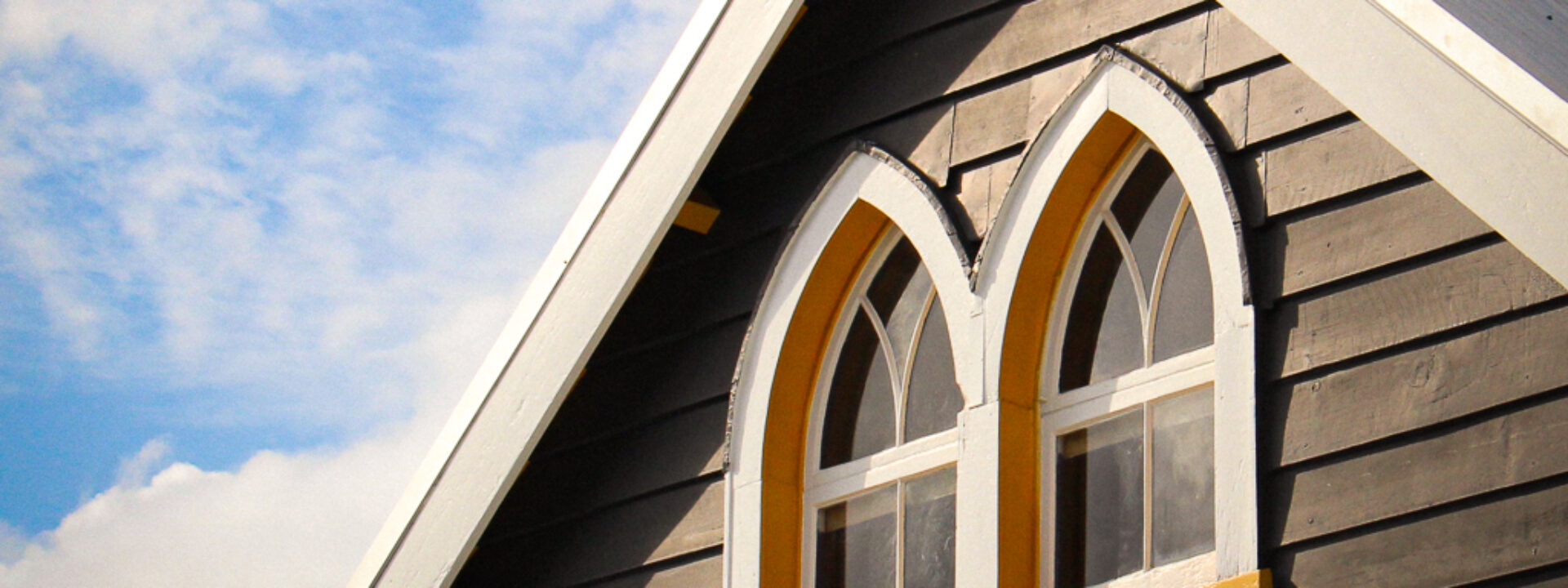 Grey weatherboard gable end with arched Gothic-style windows and yellow surrounds against blue sky