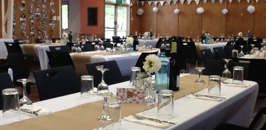 Timber hall interior set with long white-clothed tables, black chairs, and bunting near the ceiling