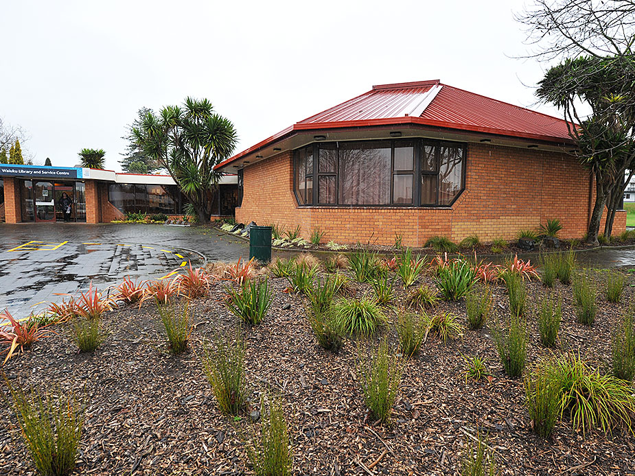 Brick community hall with red metal roof and planting beds on a wet day