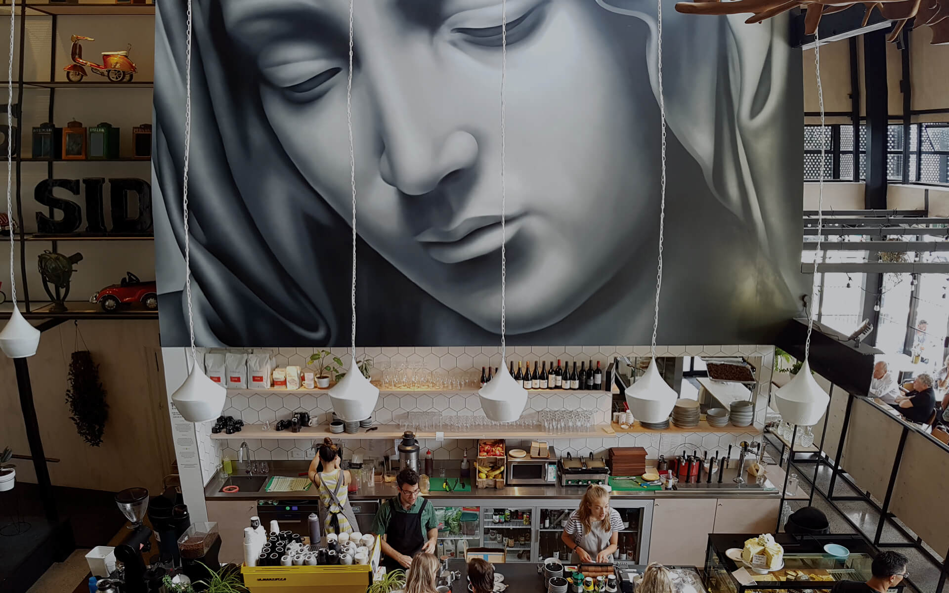Double-height cafe interior with large mural on rear wall, bar counter, hanging pendant lights, industrial shelving