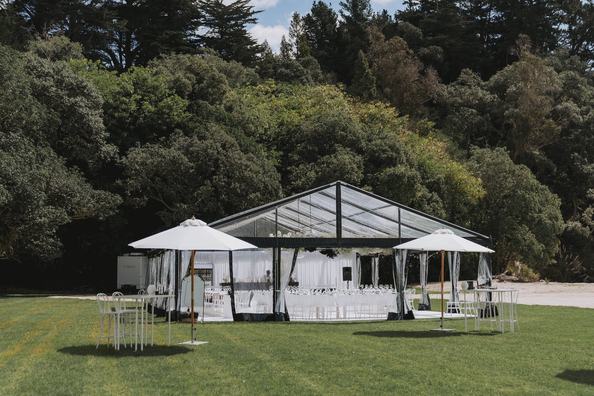 Glass and steel clear-span marquee on lawn with white chairs inside, trees and blue sky behind
