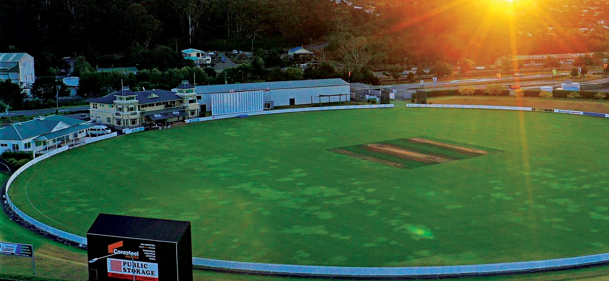 Cricket oval at dusk with a scoreboard in the foreground and surrounding hills