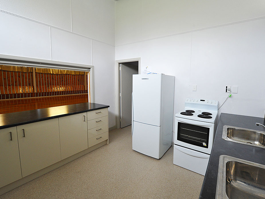 Basic kitchen with cream cabinets, dark benchtop, white fridge, and a service hatch to hall
