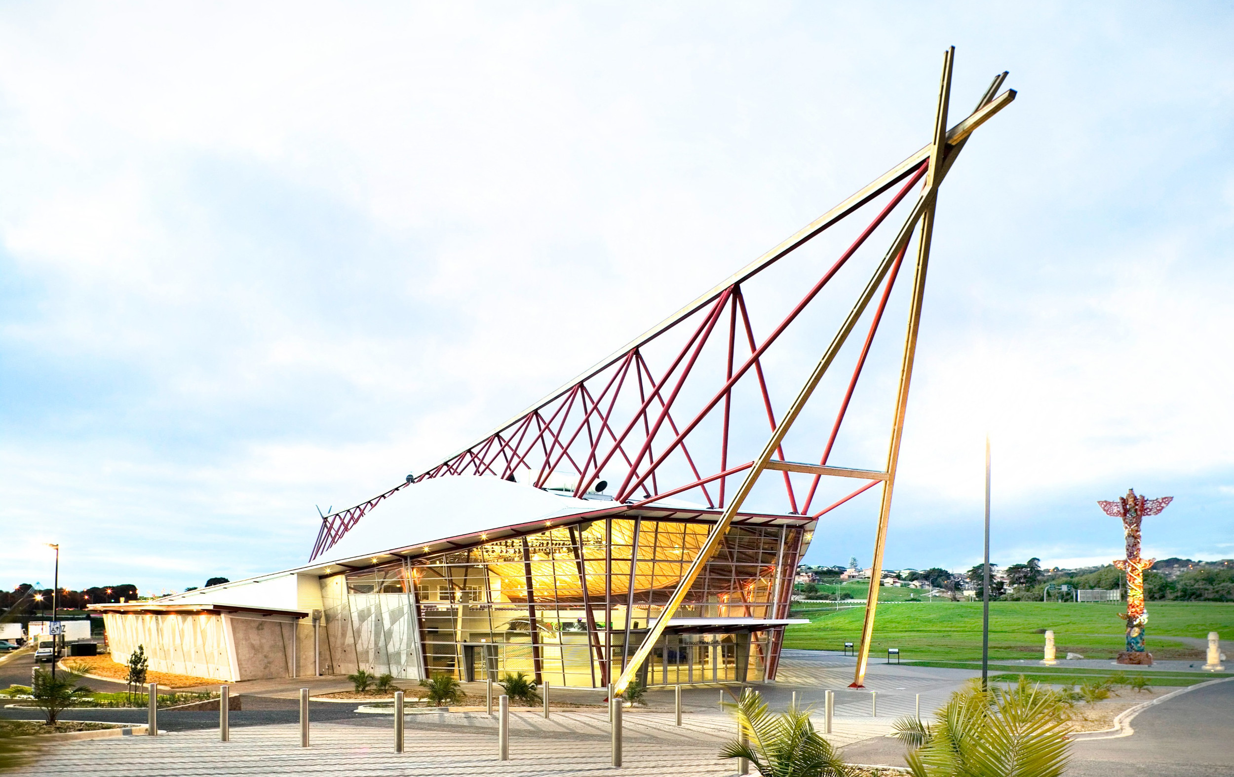 Event building exterior by day with angled red steel lattice canopy, glazed lower volume, and sculpture nearby