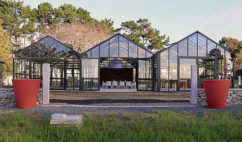 Black steel glasshouse exterior with white chairs visible inside, large planters flanking entrance