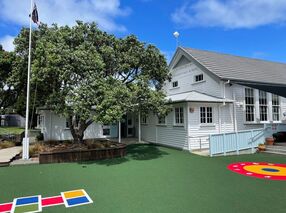 White timber school building with flagpole, trees, and playground markings