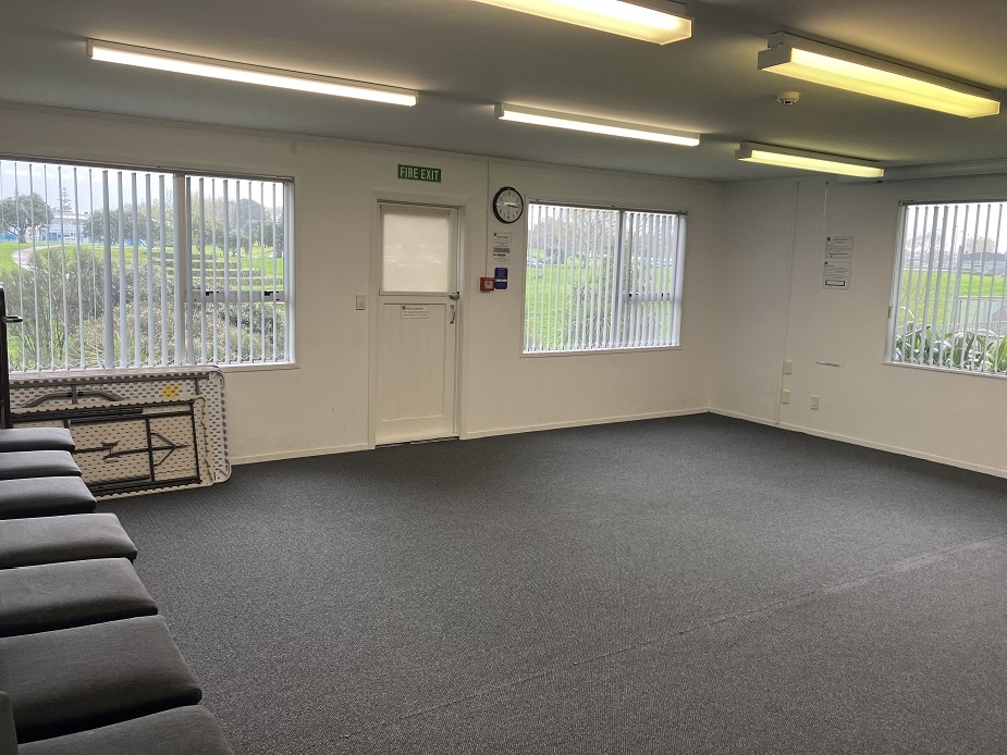 Plain carpeted room with fire exit door, two windows and folded cushions along one wall