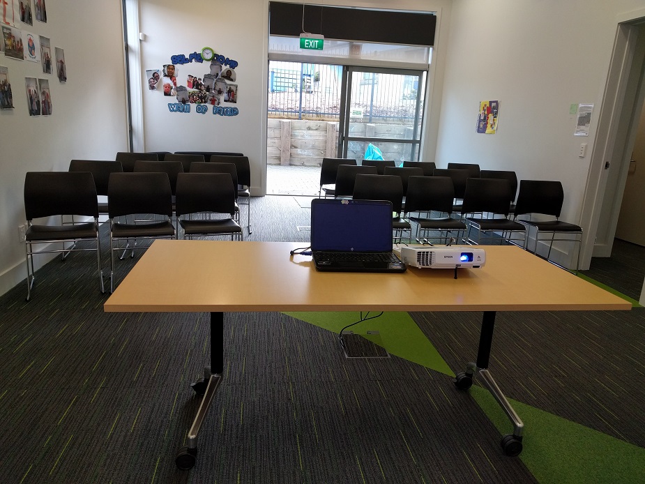 Meeting room with rows of chairs and table with projector, exit door visible