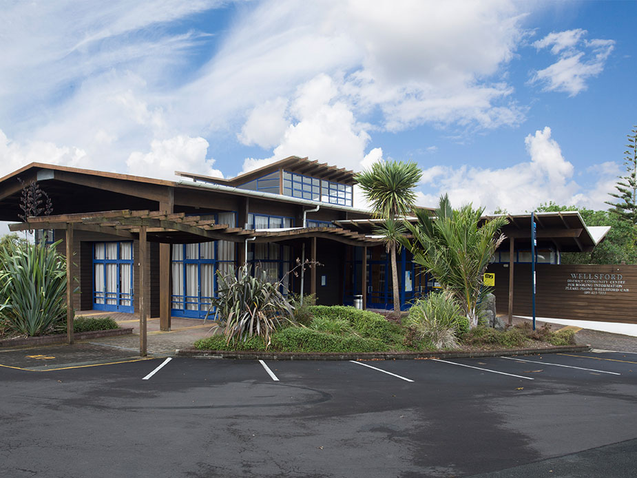 Low-rise timber and dark-stained building with pergola canopy and planted entry, car park