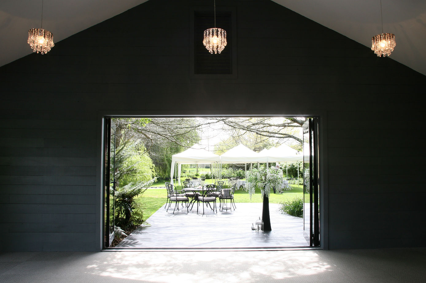A dark barn-style interior with three crystal chandeliers and open bifold doors onto a deck, garden and white marquee