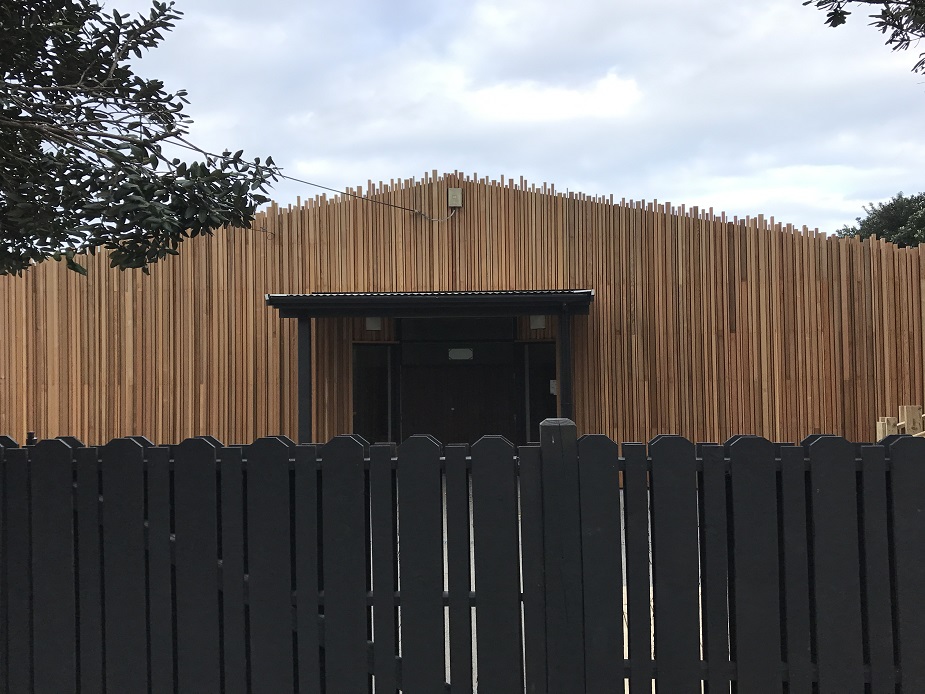 Low single-storey timber-clad building with vertical battens and jagged roofline, dark fence in foreground
