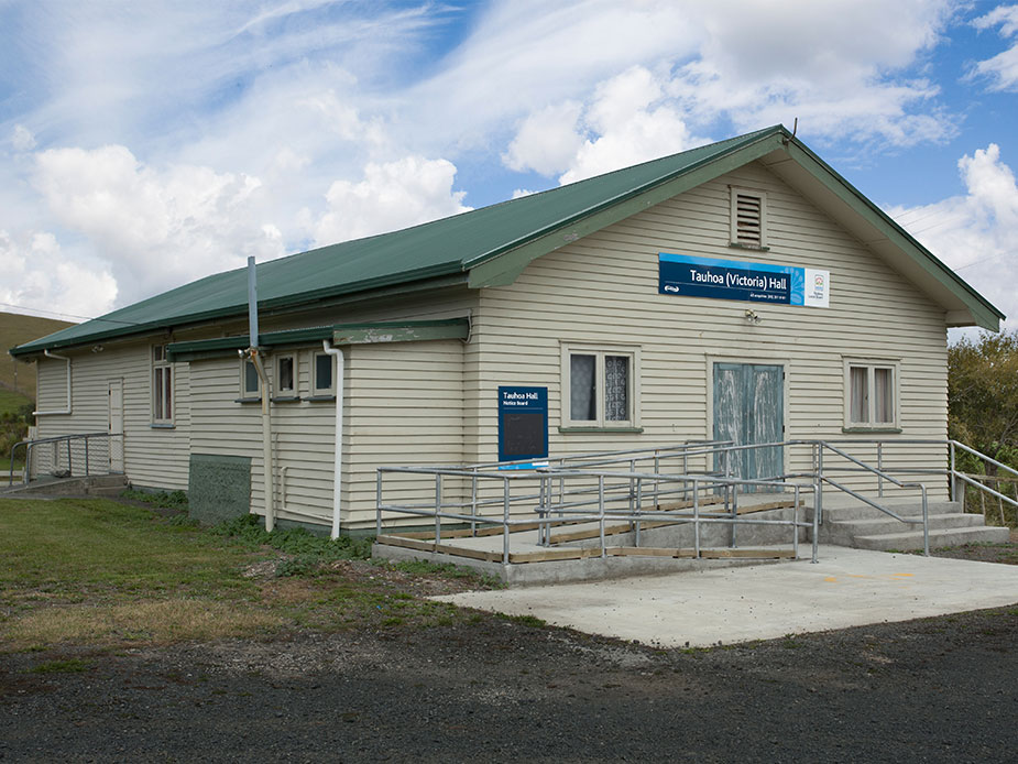 Cream weatherboard hall with green iron roof, accessible ramp, and open farmland behind