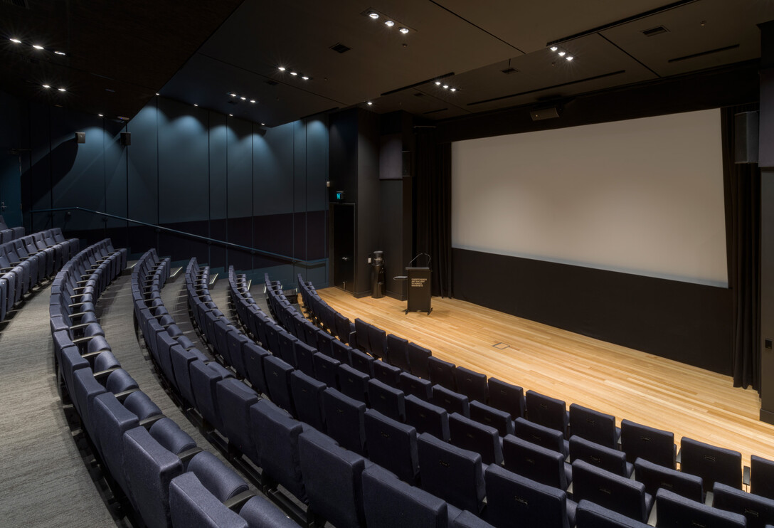 Dark blue-walled auditorium with curved rows of blue upholstered seats facing a timber-floored platform with projection screen