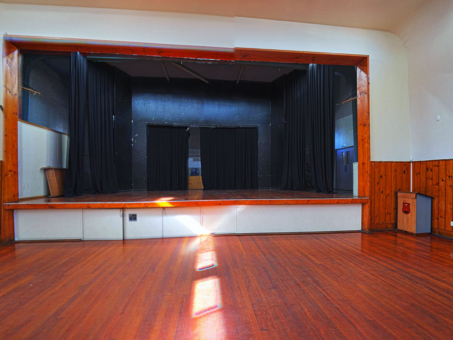 Hall interior showing raised stage with black curtains and polished timber floor