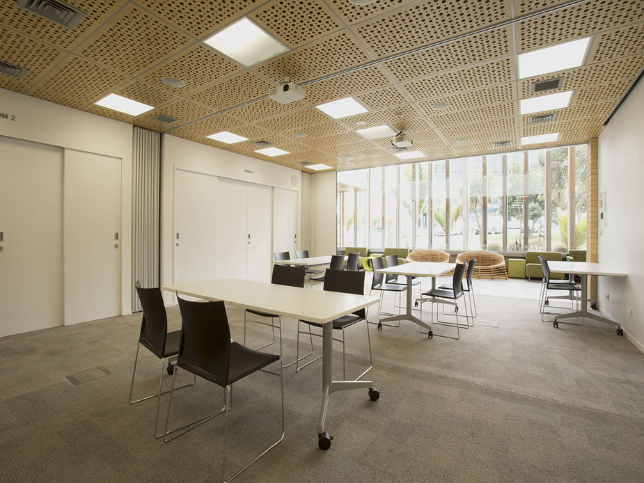 Light-filled room with perforated timber ceiling panels, white walls, movable white tables and dark chairs