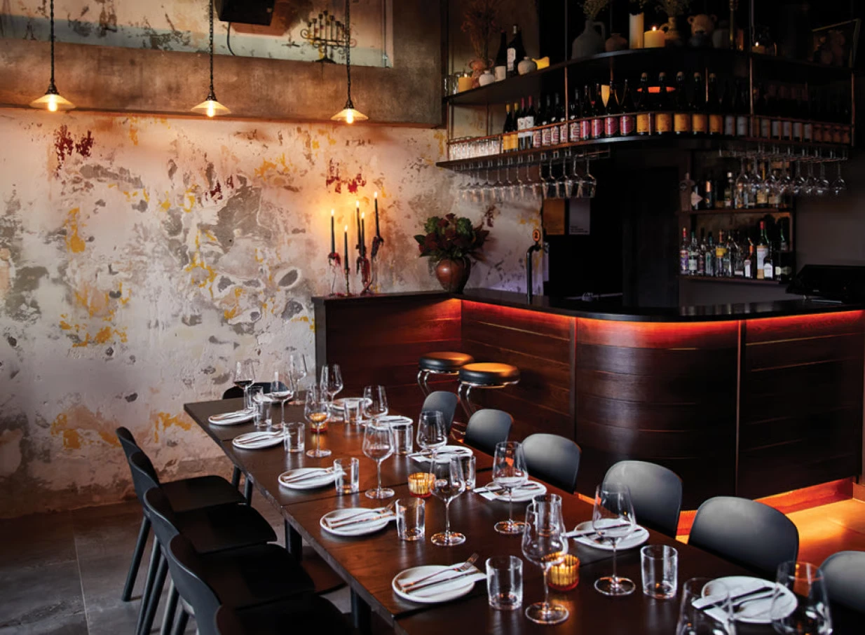 Dark dining room with distressed plaster wall, warm-lit timber bar, pendant lights, and set dining table