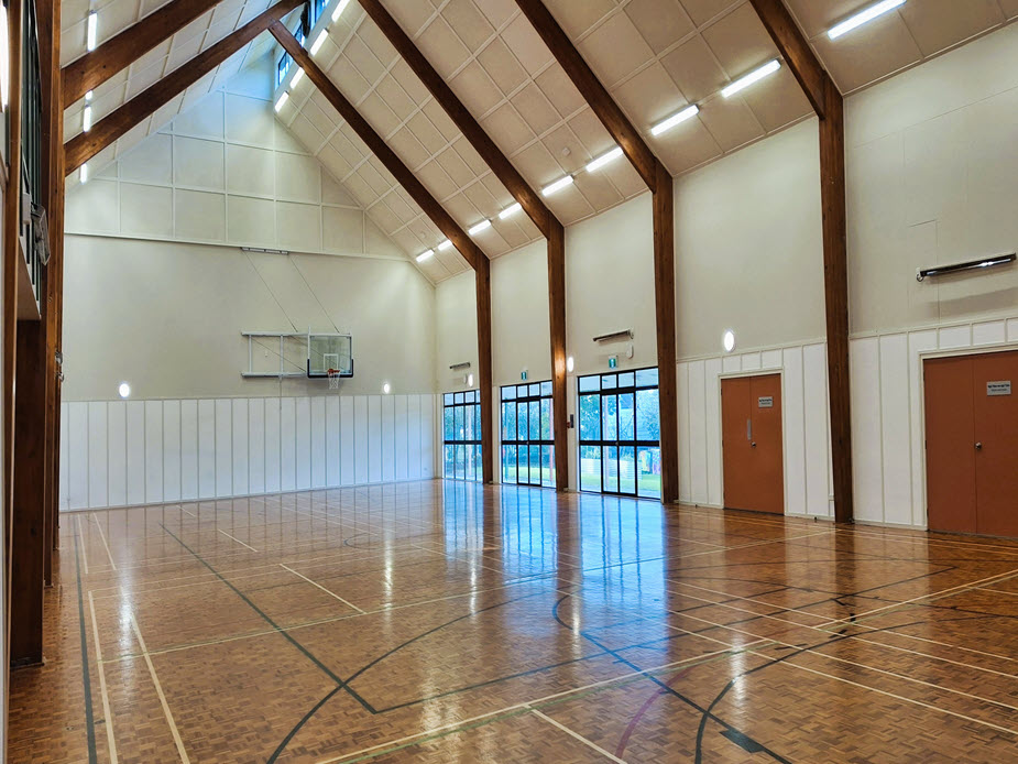 Large sports hall with vaulted timber-beam ceiling, polished timber floor, and basketball hoop