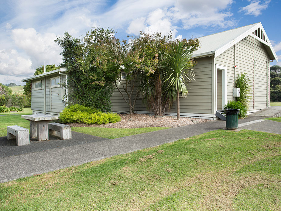 Small weatherboard hall with green gabled roof set in grass with trees behind
