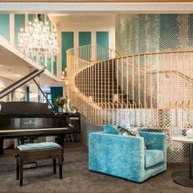 Ornate hotel lobby with grand piano, curved staircase, crystal chandelier, and teal velvet armchair