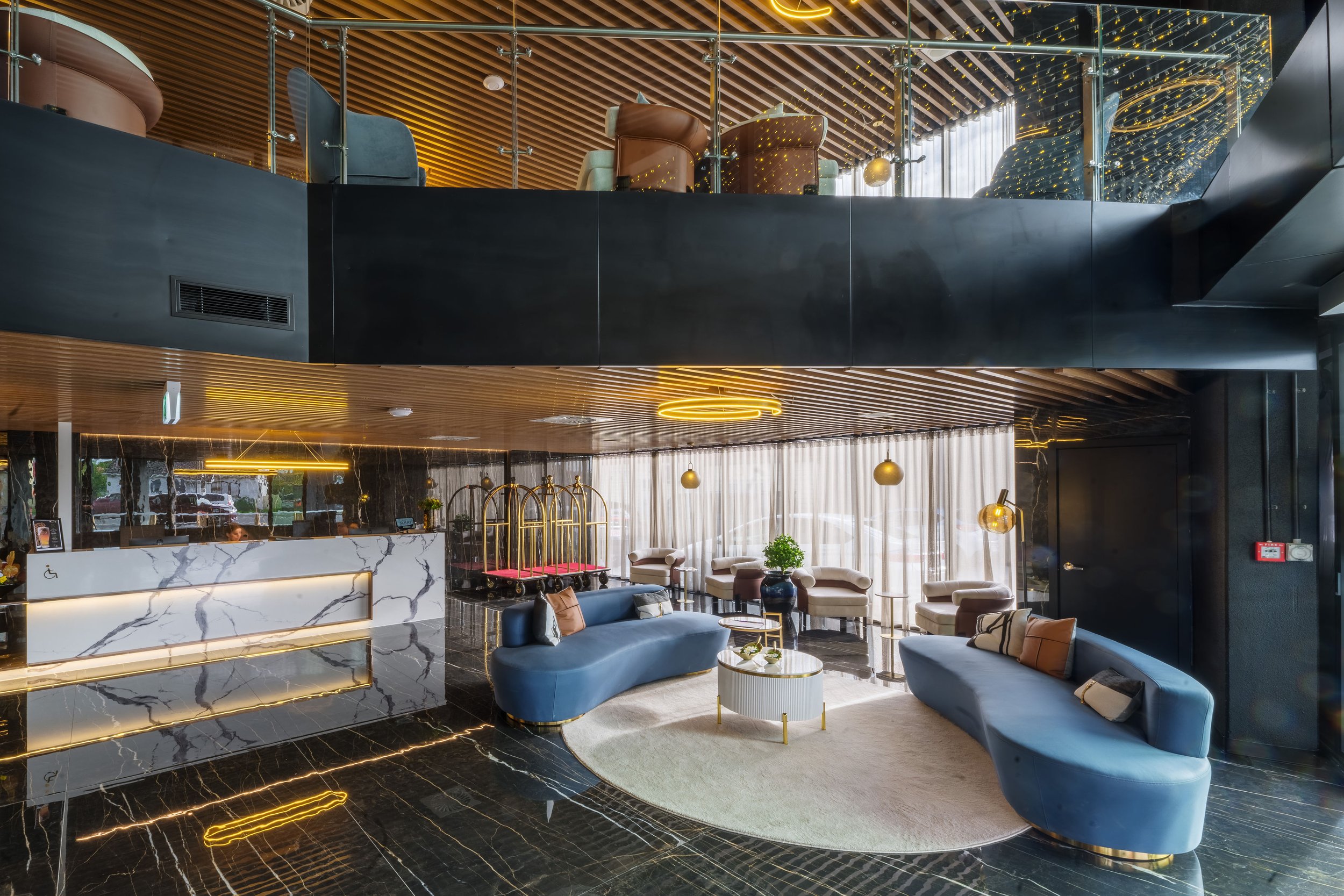 Dark marble hotel lobby with curved blue sofas, mezzanine above, timber slatted ceiling, and gold pendant lights