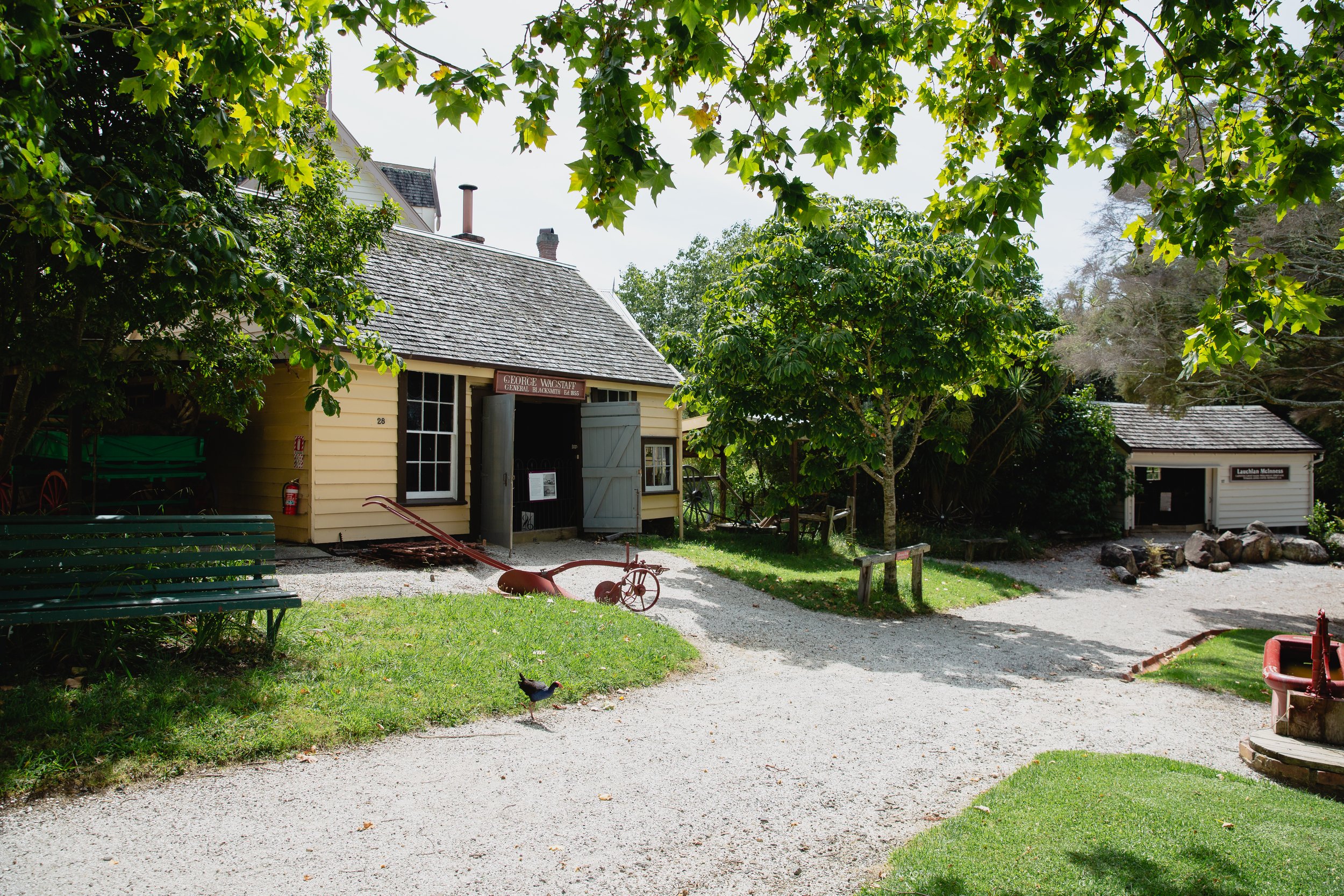 19th-century village buildings amid trees with gravel paths and farm equipment