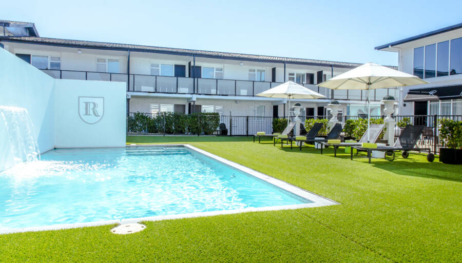 White two-storey hotel building with pool, sun loungers, umbrellas, and manicured lawn