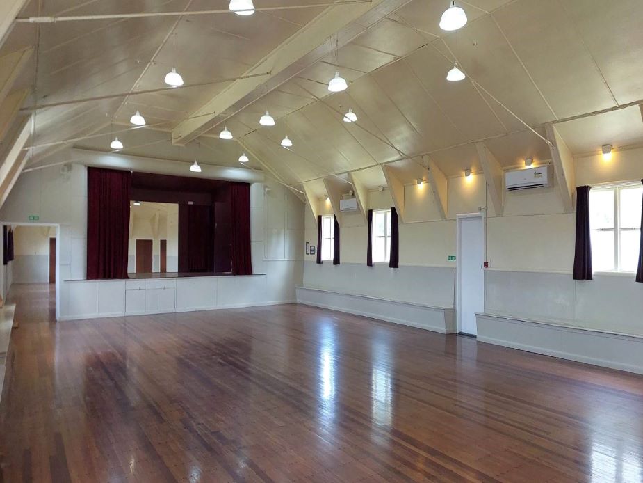 Timber-floored hall interior with arched ceiling, pendant lights, red stage curtain, and tall sash windows