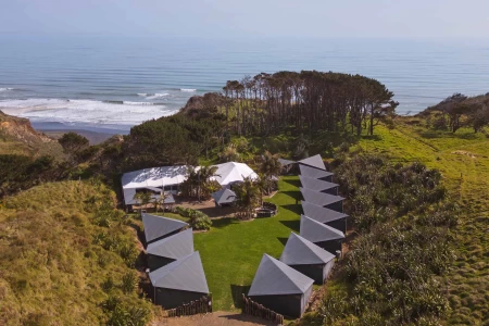 Aerial view of canvas tent village with large marquee on grassed coastal property, trees and ocean visible