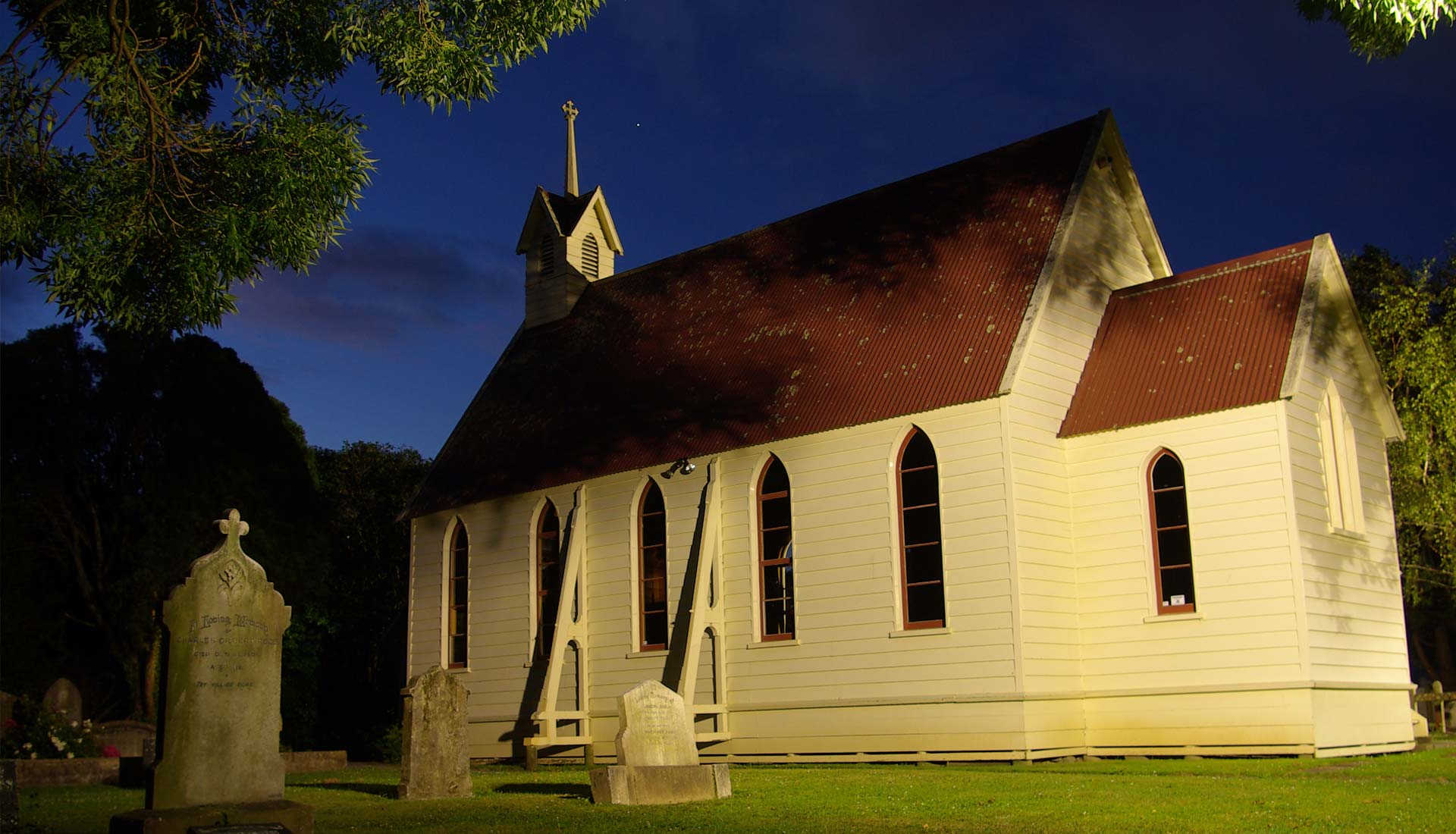 Small white timber church illuminated at night with historic graves in foreground