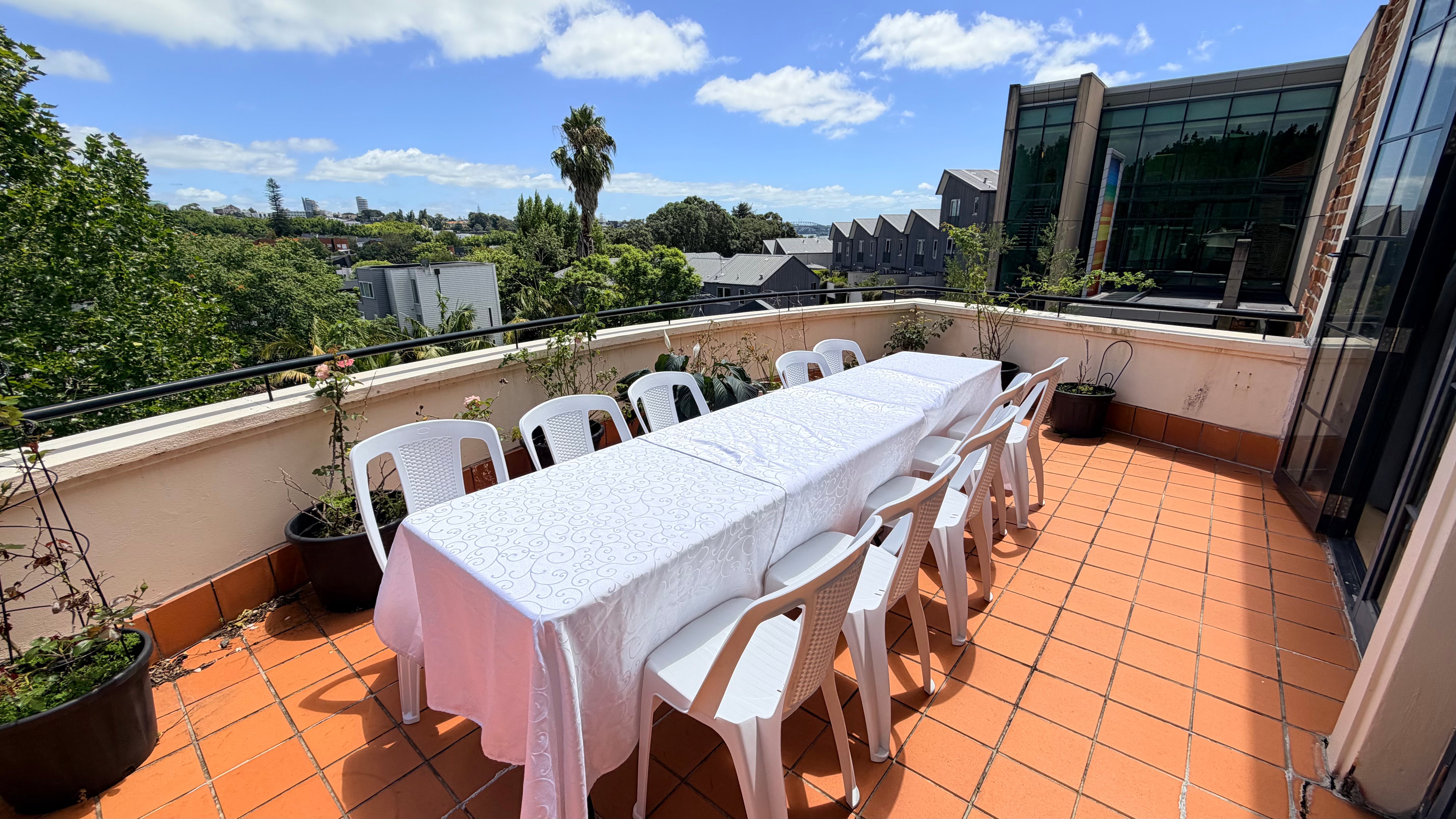 Rooftop terrace with long white-clothed table, white chairs, and city views in background