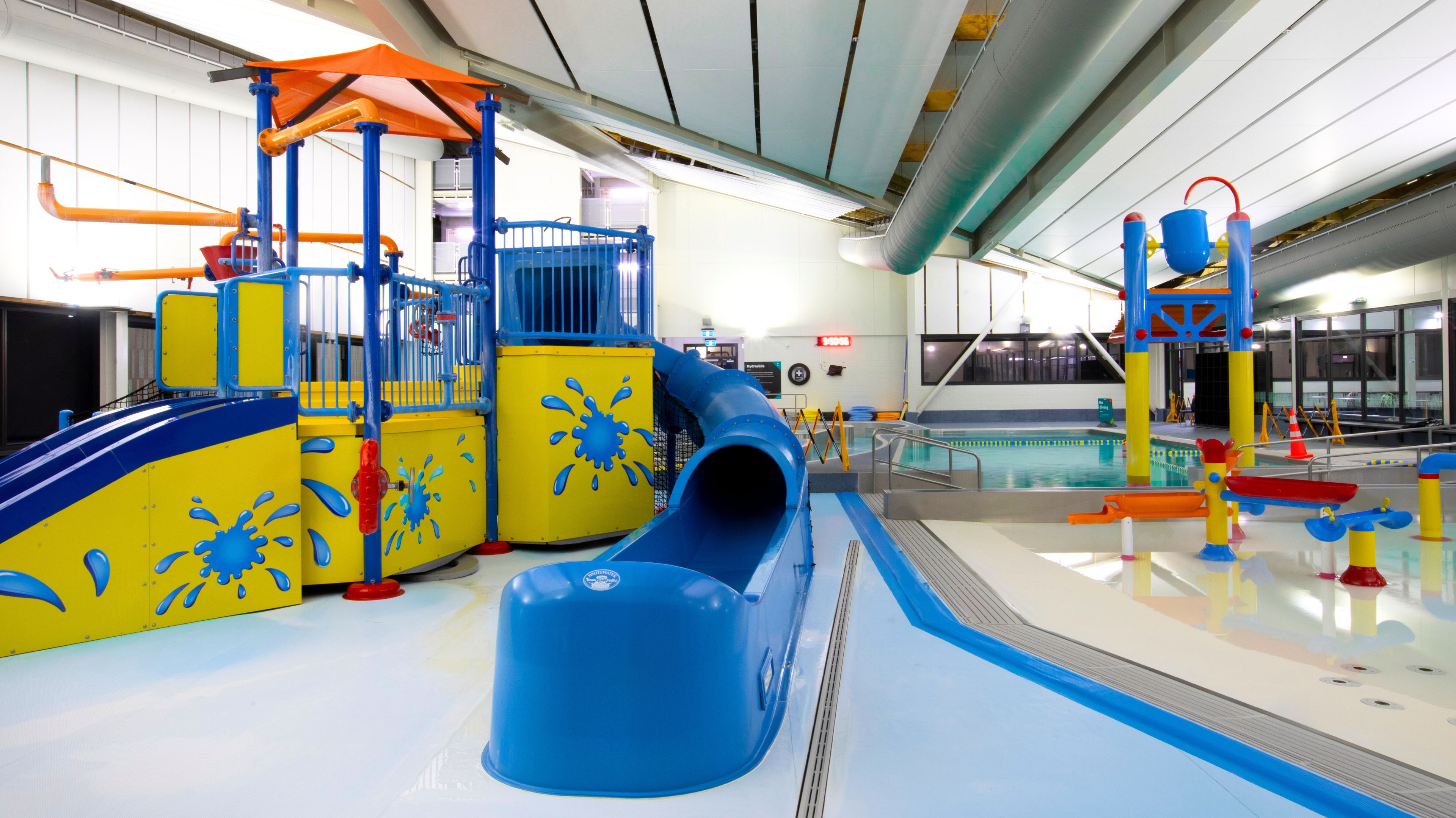 Indoor water play area with coloured slide and play structures beside a pool