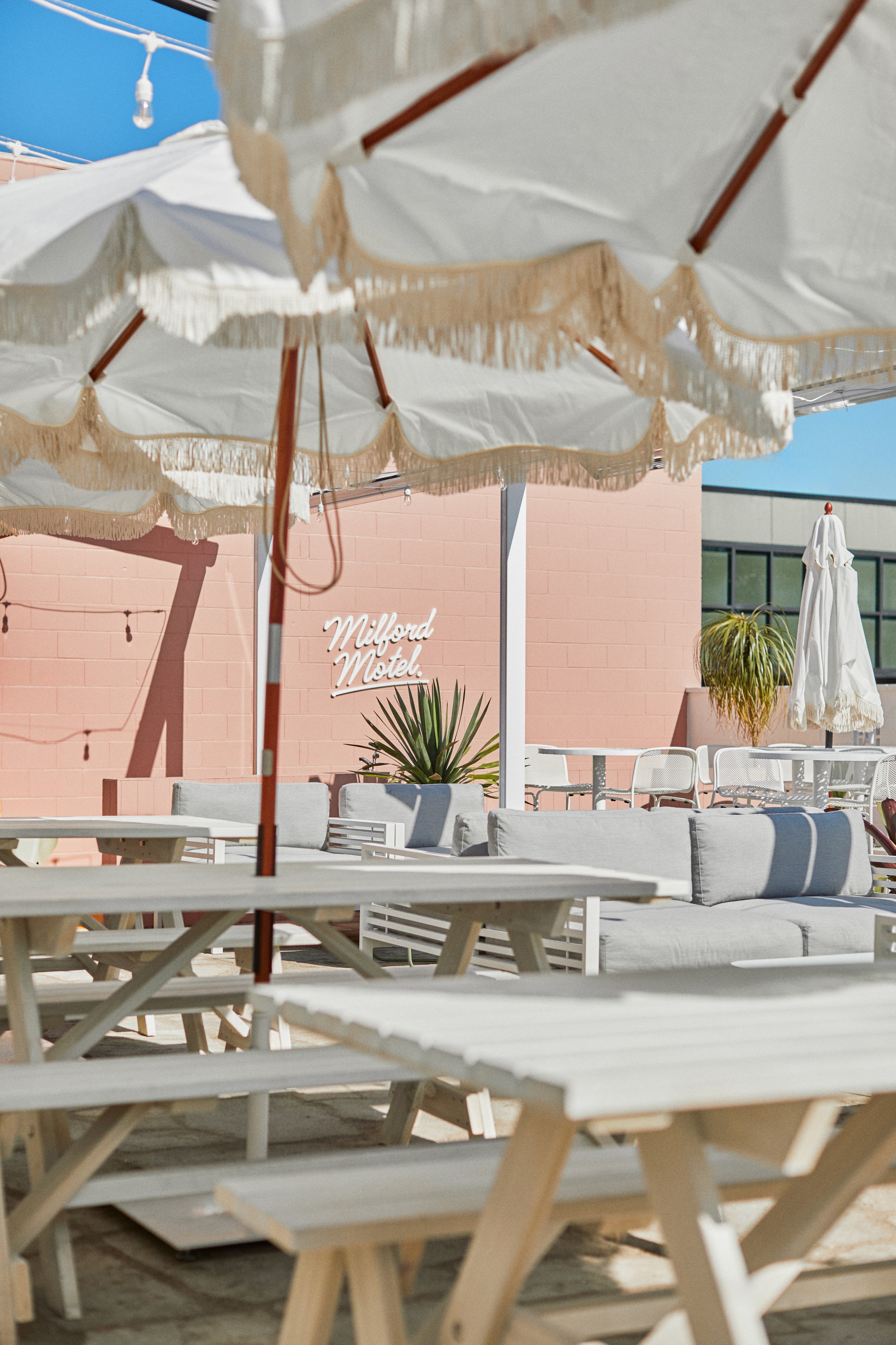 Outdoor seating area with fringe umbrellas, bench tables and neon sign on pink wall