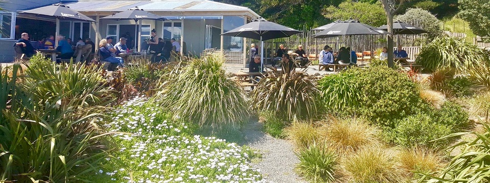 An outdoor seating area with dark umbrellas and picnic tables among native flax beside the hotel