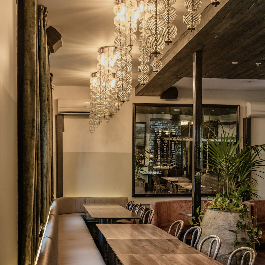 Warm dining room with globe chandeliers, timber ceiling, banquette seating, and wine display beyond glass partition