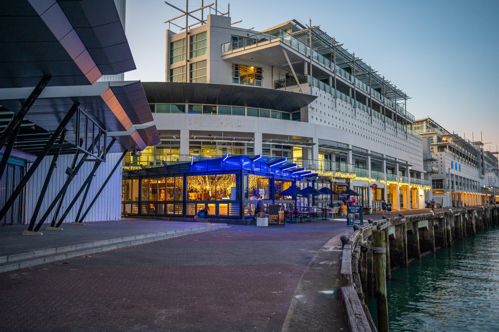 Waterfront bar and dining pavilion with blue neon canopy, dusk, harbour backdrop