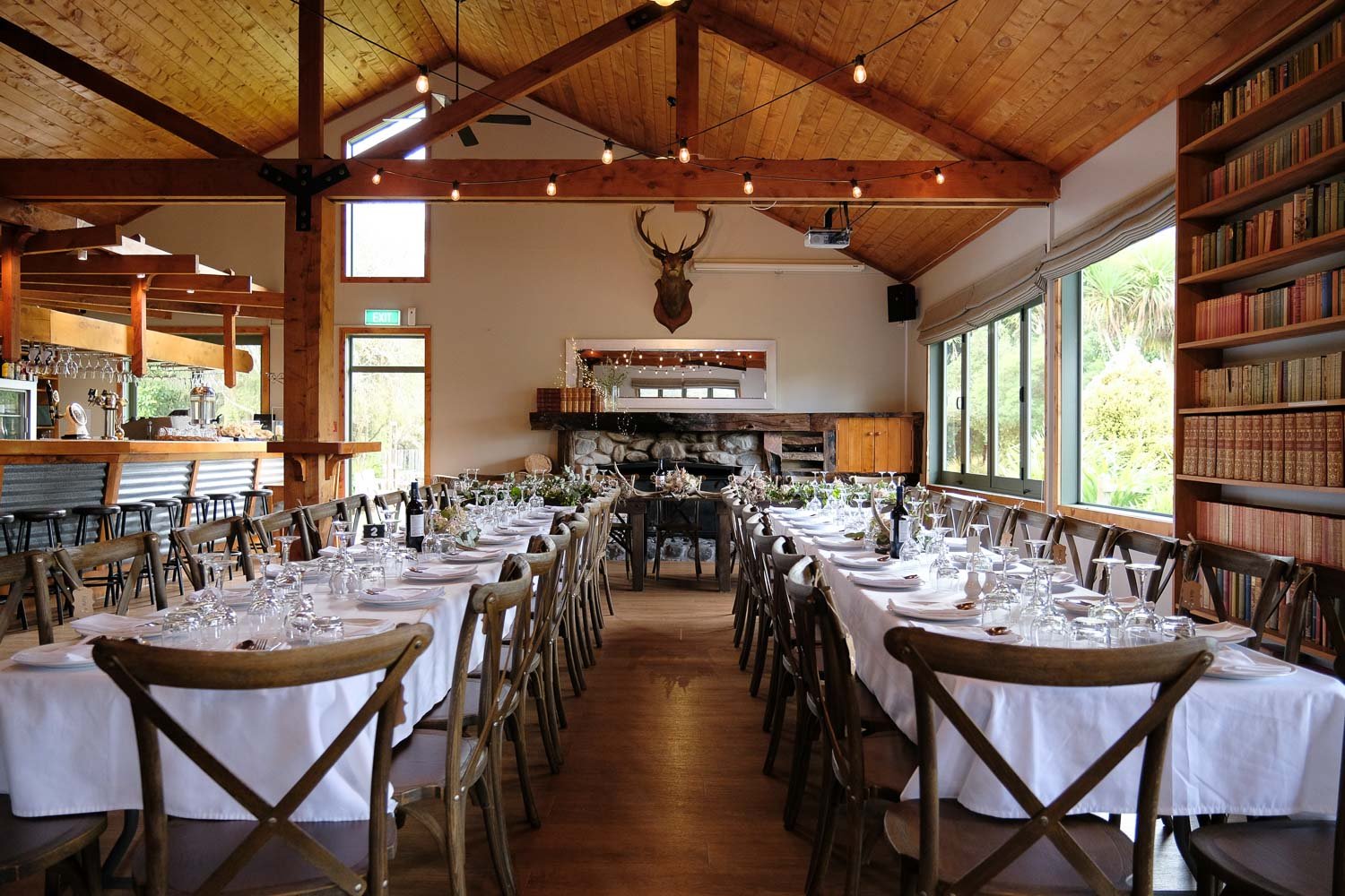 High-stud timber-framed reception hall with long banquet tables, a stone fireplace and string lights