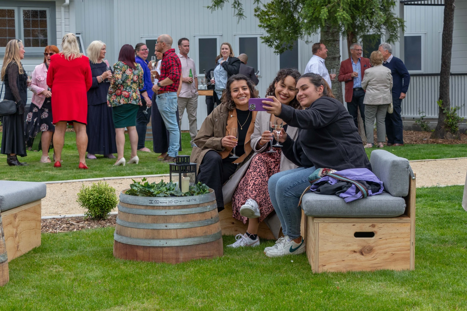 Guests gathered on a lawn beside a white weatherboard building with outdoor barrel furniture