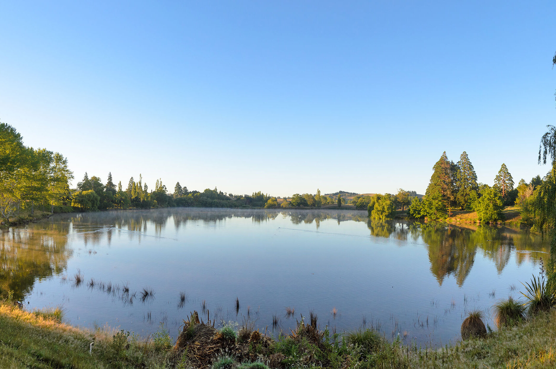 Still lake surrounded by trees reflected in calm water, open countryside beyond under clear sky