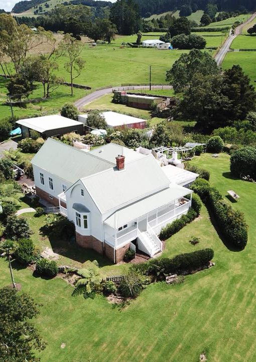 White two-storey homestead with verandah and metal roof, aerial view over green lawns