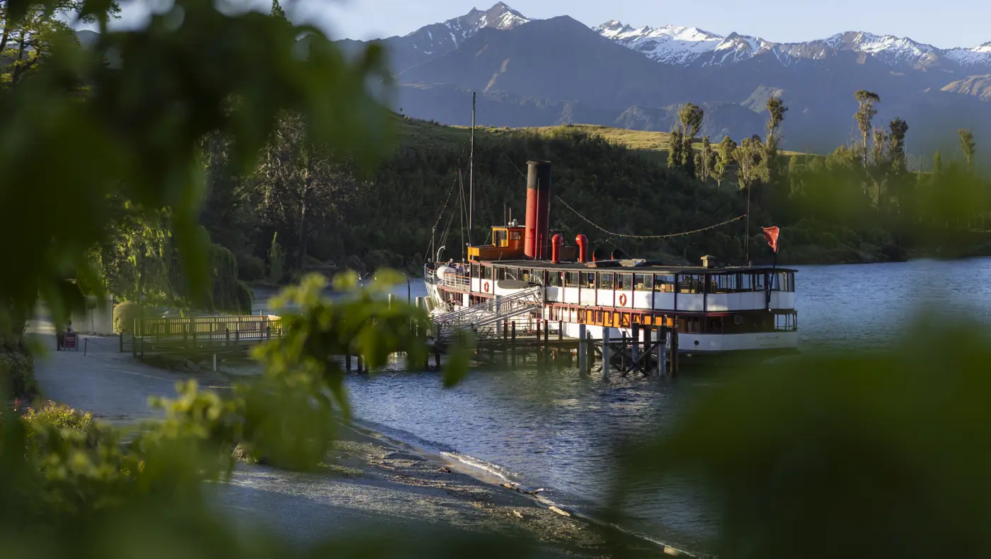 Vintage steamship moored at a timber wharf with trees and snow-capped mountains beyond