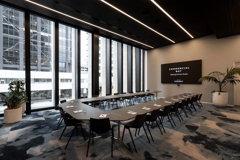 Modern boardroom with U-shaped table, dark chairs, floor-to-ceiling windows with city high-rise views and wall-mounted screen