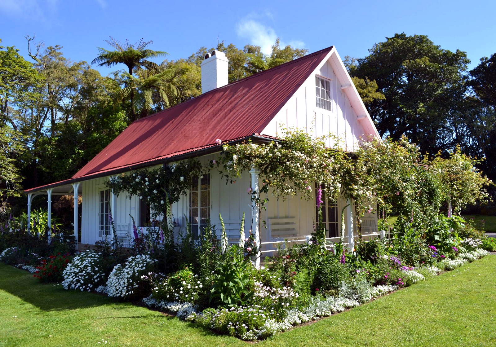 White cottage with red roof surrounded by cottage garden in full bloom