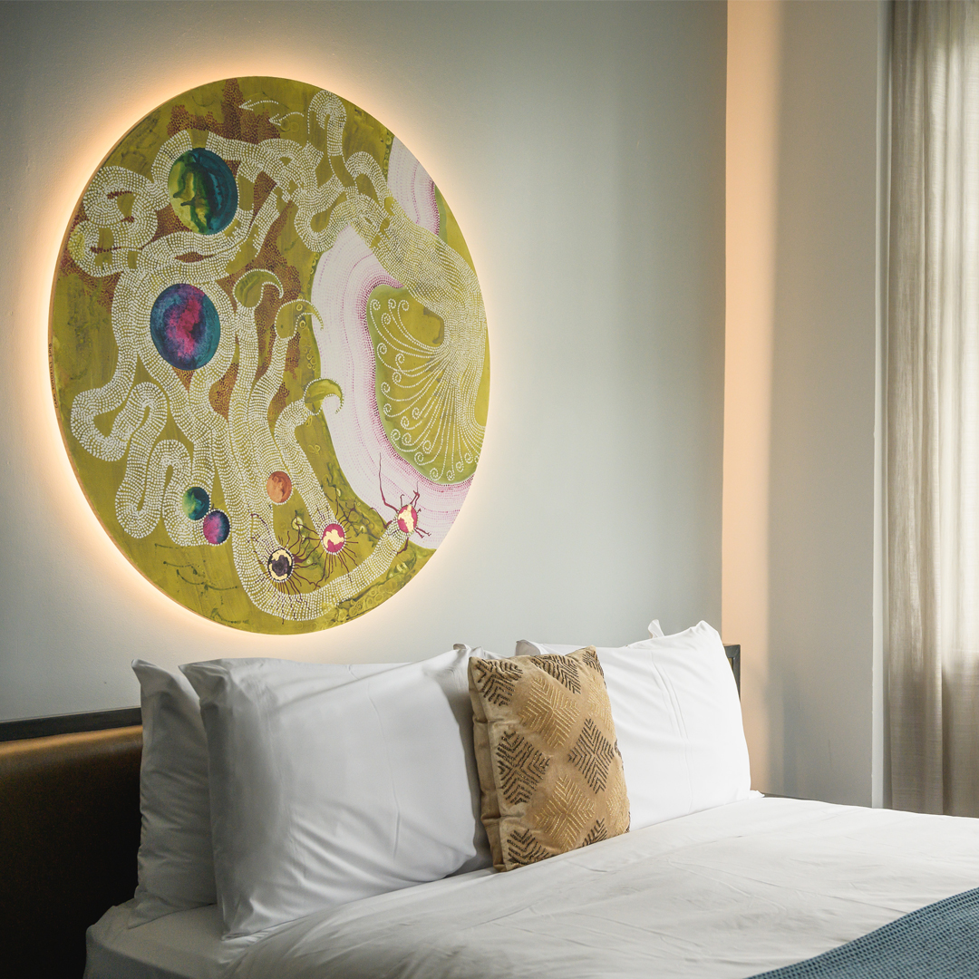 White hotel room with double bed, large circular backlit artwork above headboard, and sheer curtained window