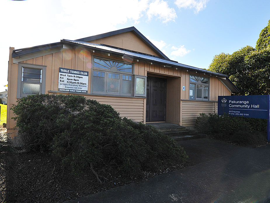 Timber-clad community hall with dark door, signage and overgrown shrubs