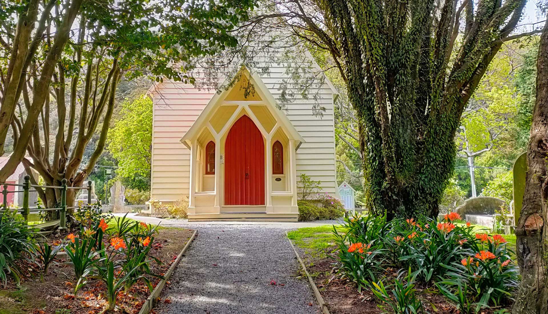 Small cream timber church with red roof and gardens in foreground