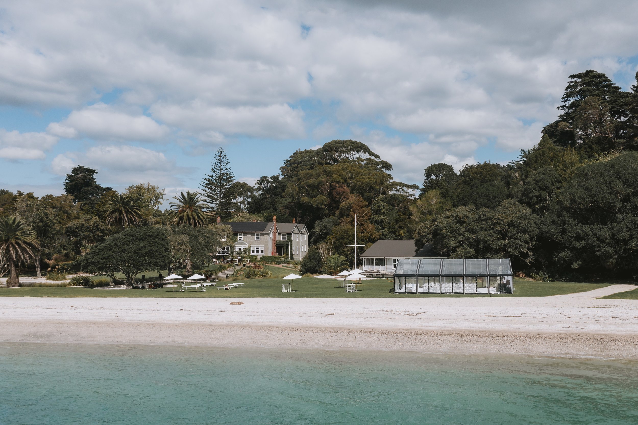 Stone homestead and glass marquee on lawn beside sandy beach with trees behind, viewed from water