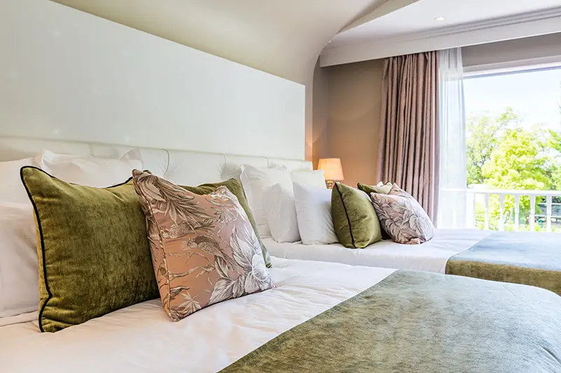Twin hotel room with green and pink cushions, white bedding, balcony with tree view